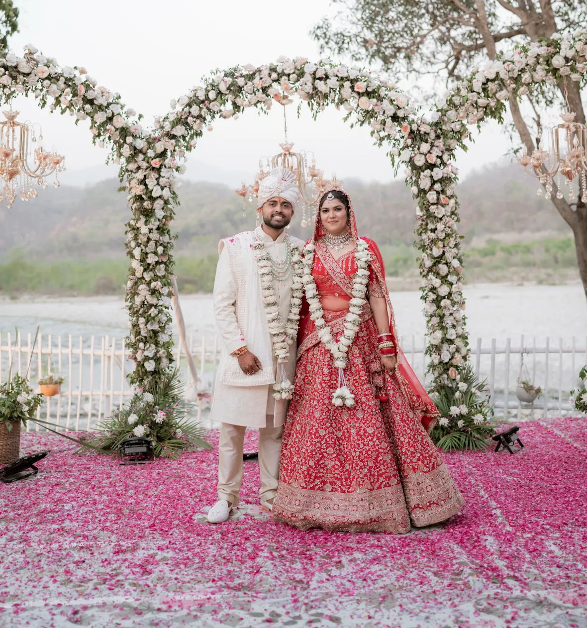 The couple in garlands beneath a white floral arch by the river