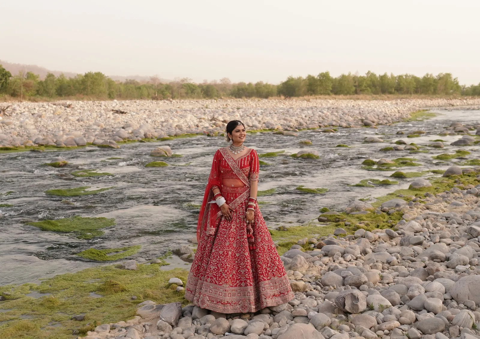 A bride in a red lehenga standing alone on the rocky bed of a river at dusk